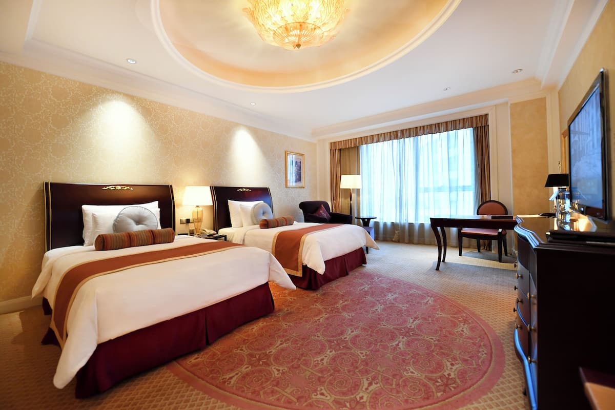 A hotel room with two beds, a desk, a chair, a TV, and a large window with curtains.