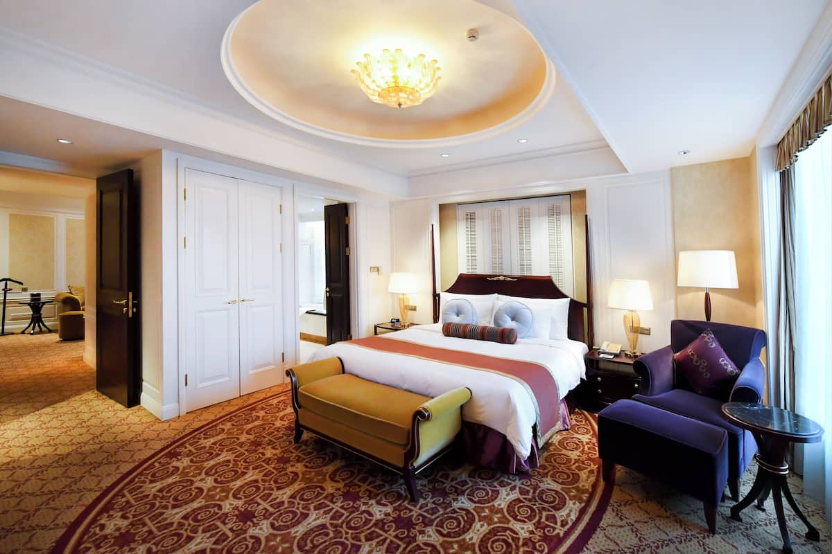 A hotel room with a large bed, a sitting area with a chair and ottoman, a small table, and a chandelier.