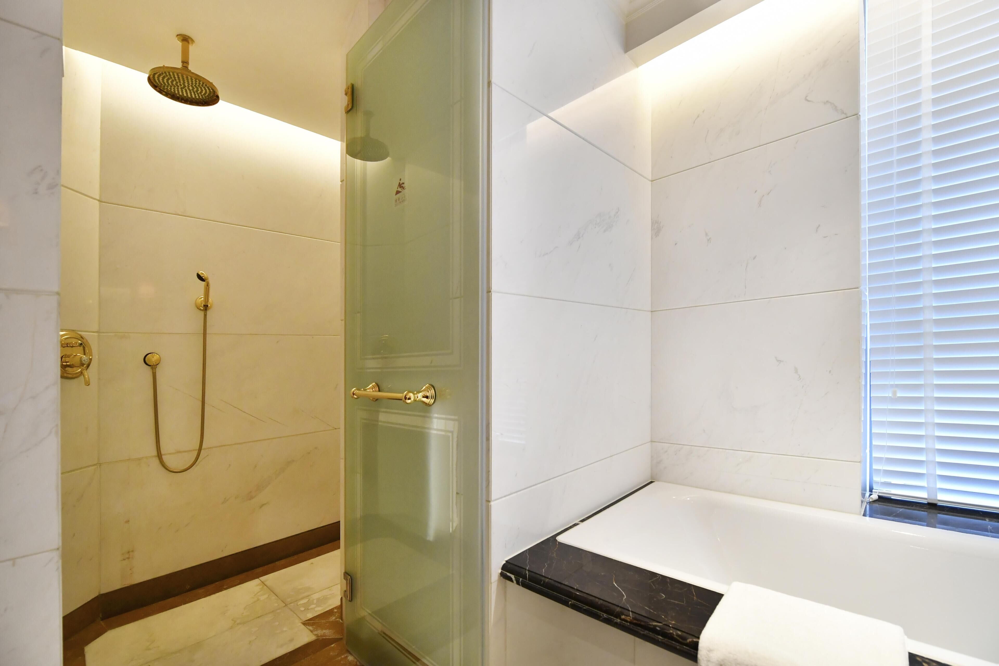 A modern bathroom with a glass-enclosed shower, a bathtub with a black marble surround, and a window with blinds.