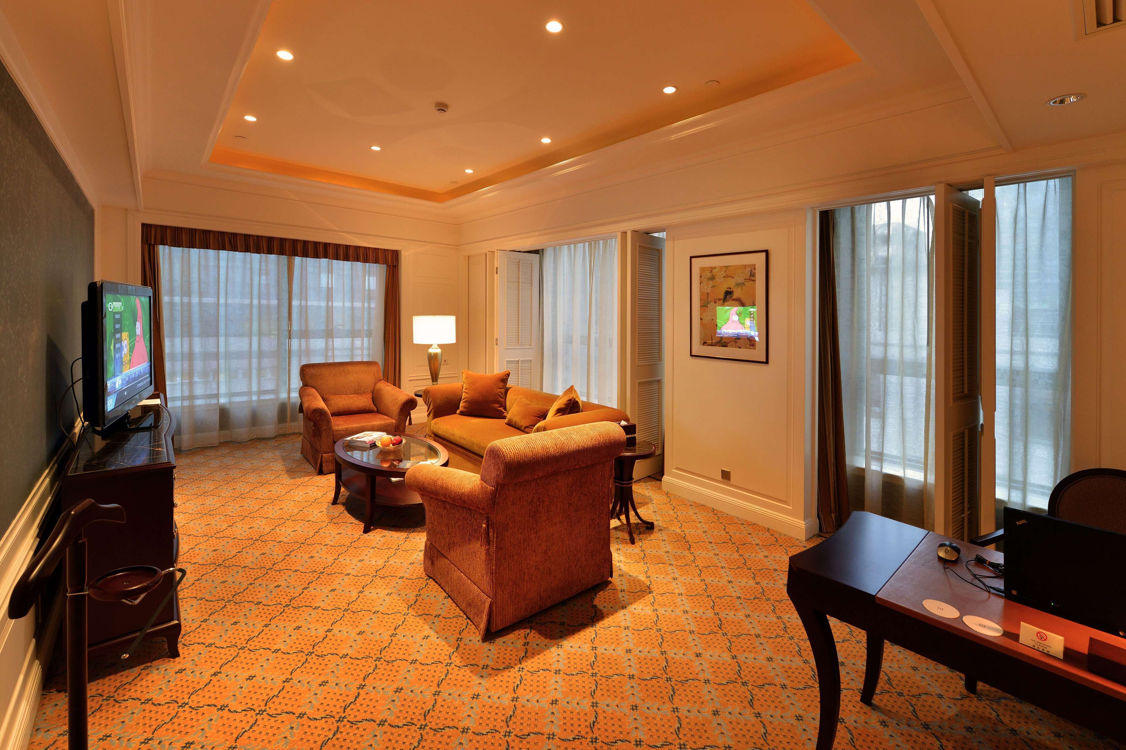 A hotel room with a sofa, armchair, TV, desk, and large windows.