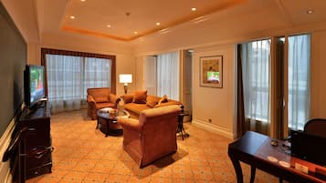 A hotel room with a sofa, armchair, TV, desk, and large windows.
