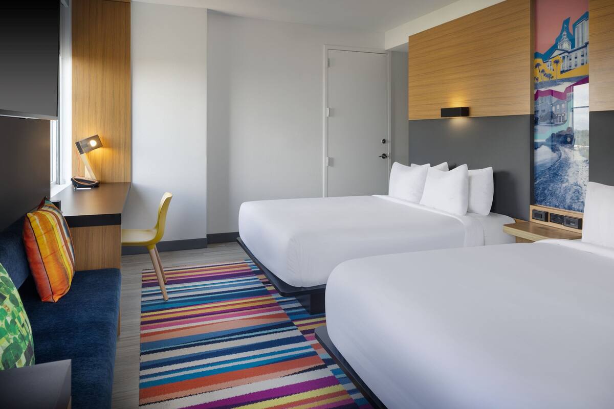 A hotel room with two beds, a desk with a lamp, a colorful striped rug, and a painting on the wall.
