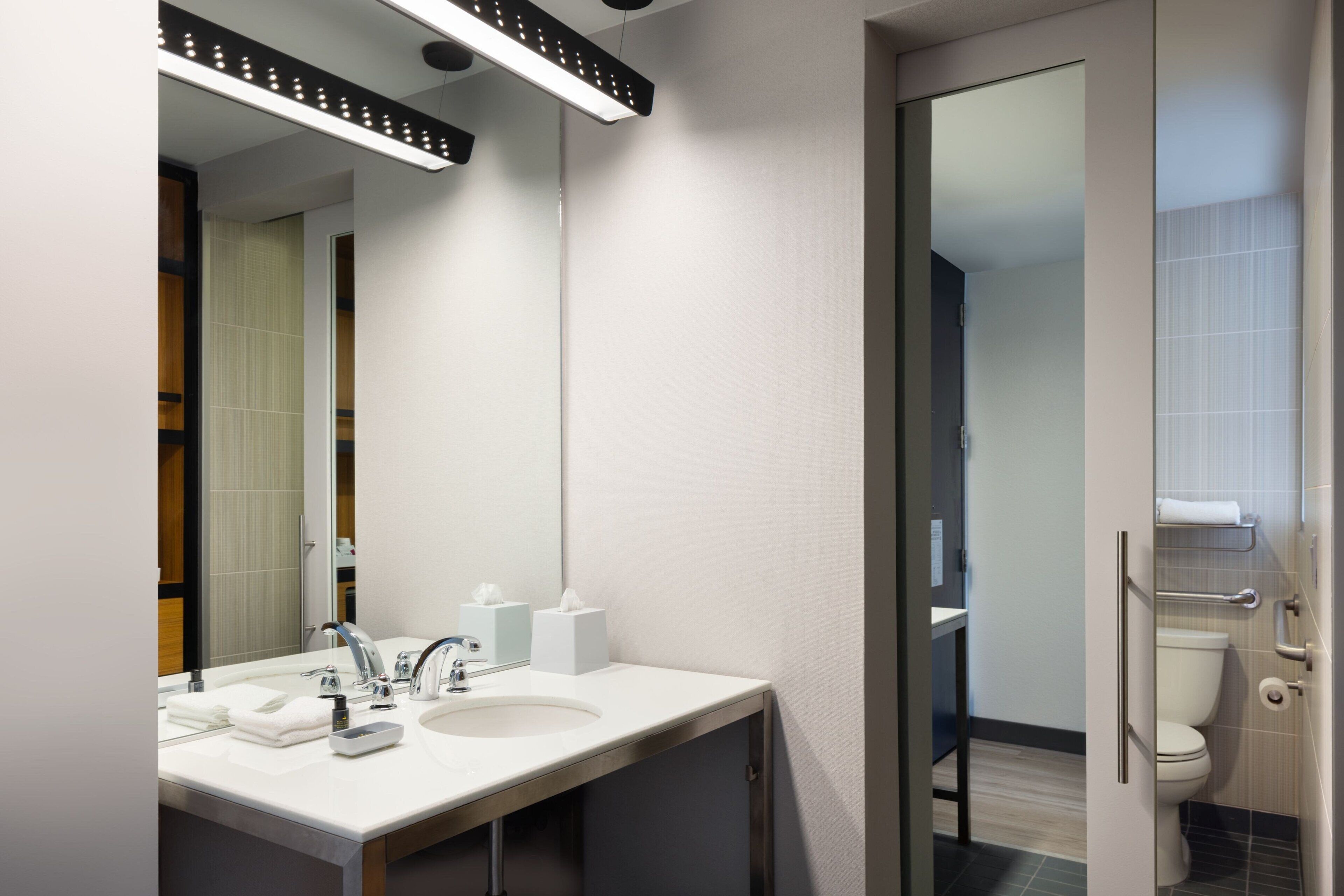 Une salle de bain moderne avec un lavabo blanc, un miroir et une porte en verre donnant accès aux toilettes.