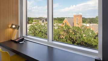 A room with a desk and chair, a window with a view of a church and surrounding buildings.