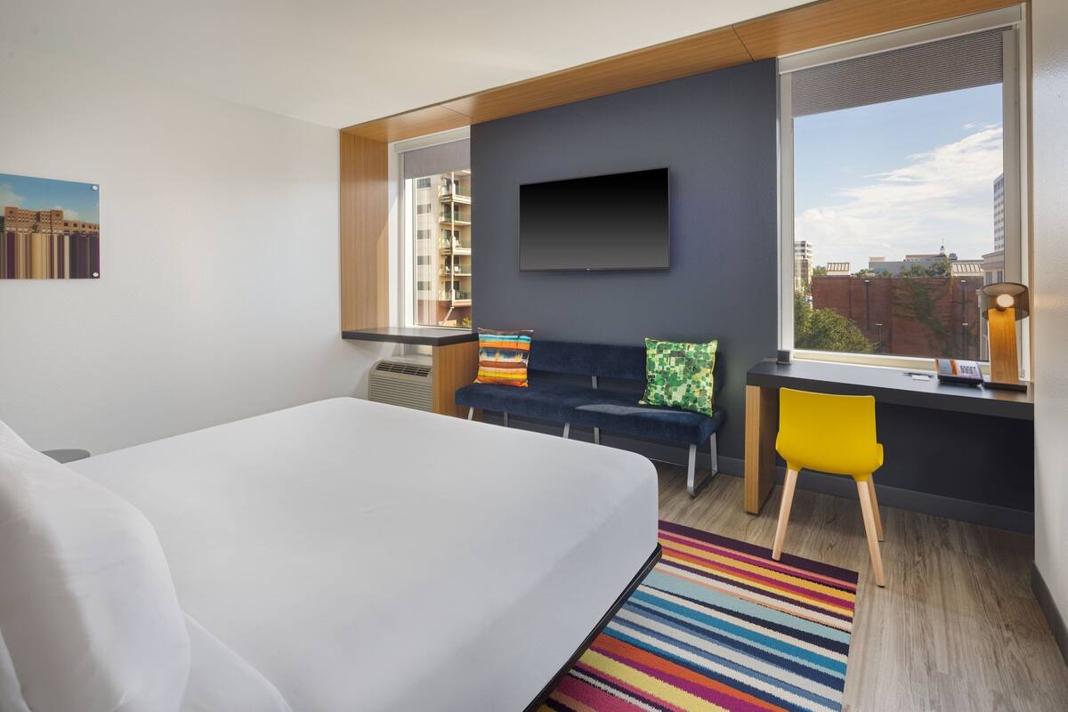 A modern hotel room with a large bed, a desk with a chair, a TV mounted on the wall, and a window with a city view.