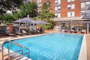 A swimming pool with lounge chairs and umbrellas, surrounded by a brick building with balconies. - Aloft by Marriott Tallahassee Downtown (Tallahassee)