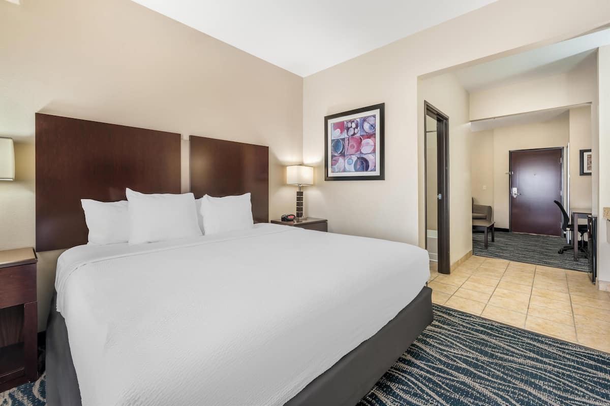 A hotel room with a large bed, a nightstand with a lamp, a desk with a chair, and a door leading to another room.