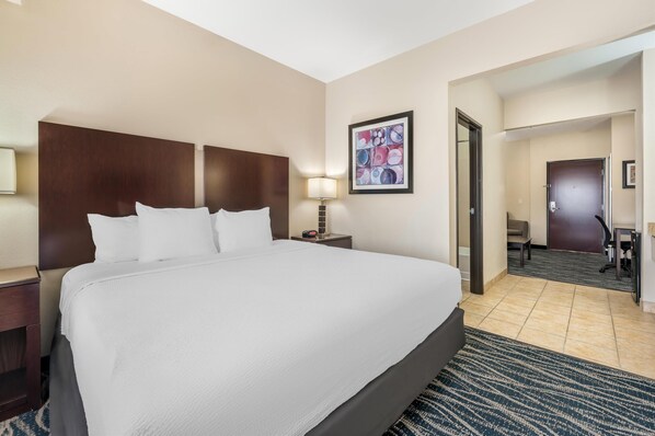 A hotel room with a large bed, a nightstand with a lamp, a desk with a chair, and a door leading to another room.