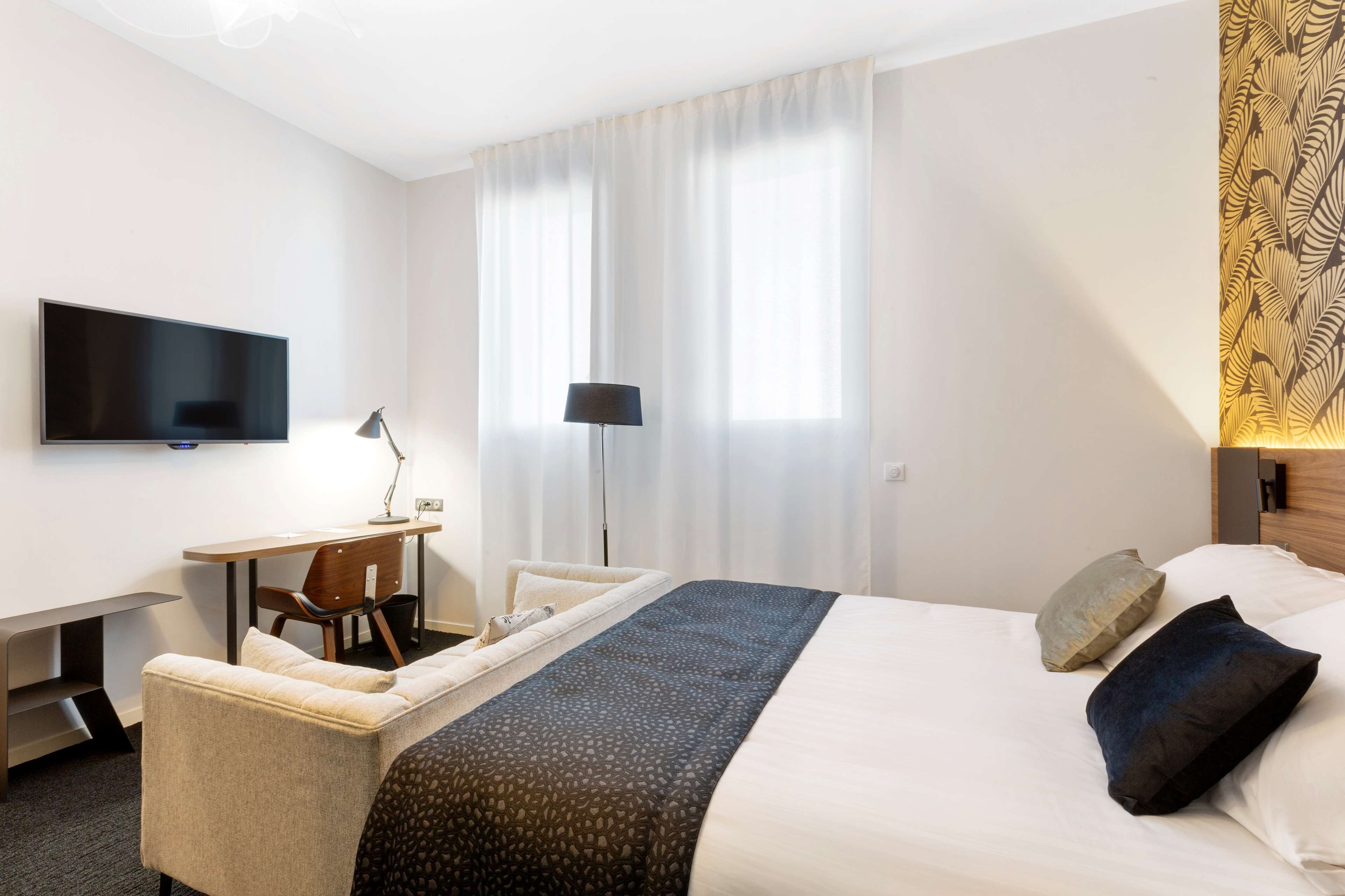Habitación de hotel con cama, escritorio, silla, televisor y ventana con cortinas.