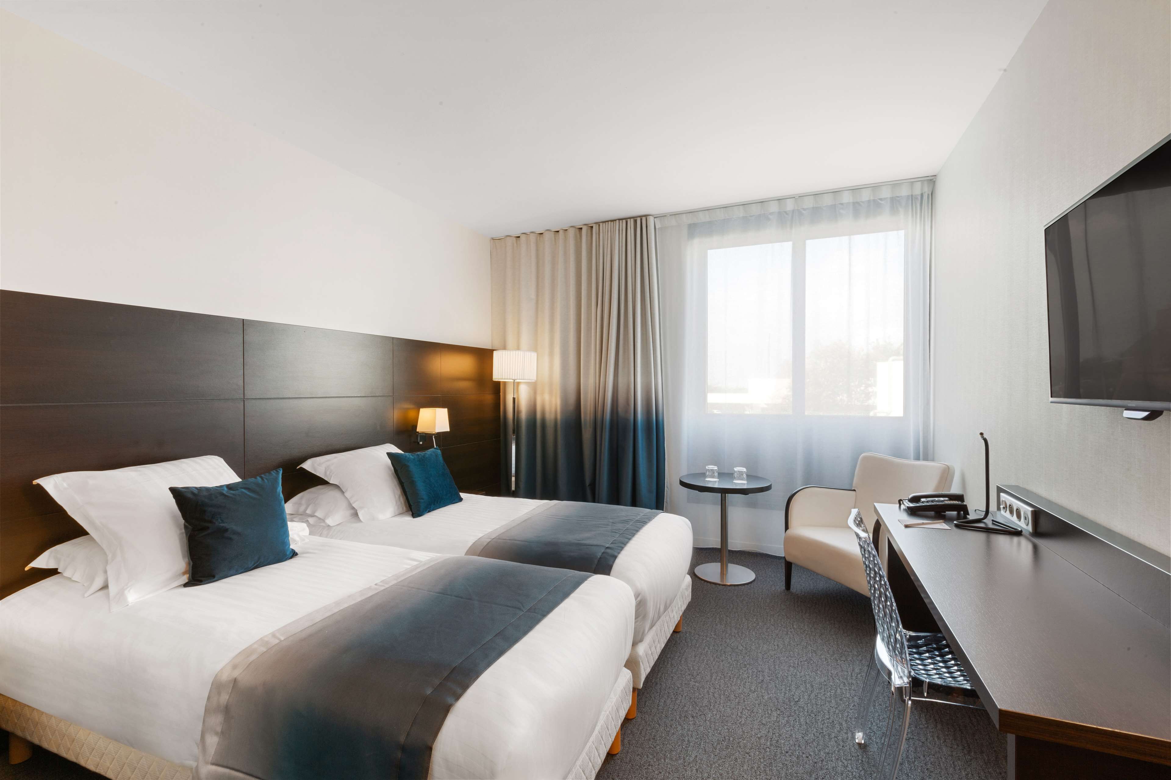 A hotel room with two beds, a desk, a chair, a TV, and a small table with a lamp.