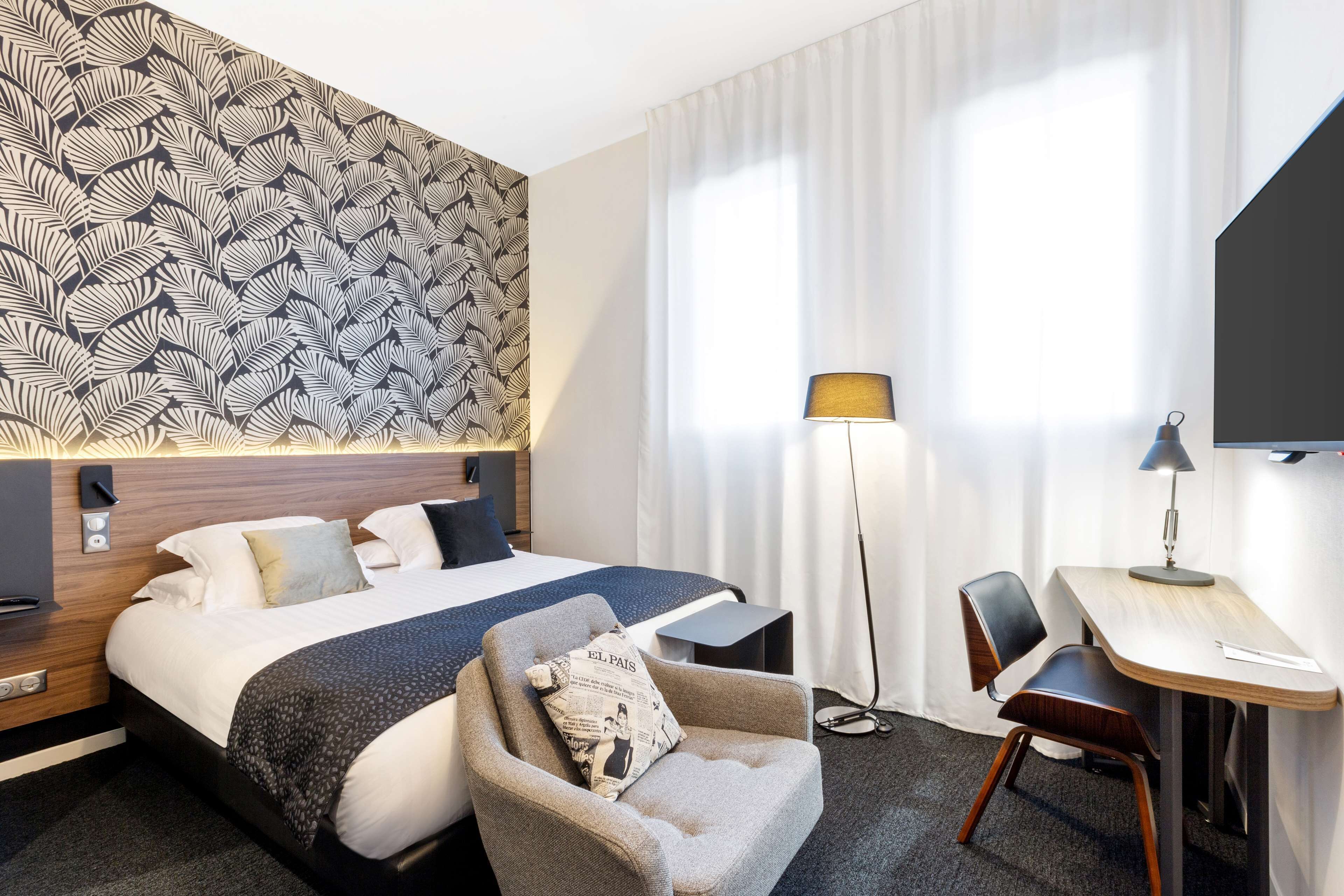 A hotel room with a bed, a desk, a chair, a lamp, and a television.