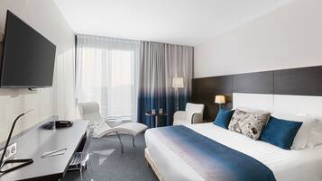 A hotel room with a large bed, a desk with a computer, a chair, a TV, and a window with curtains.