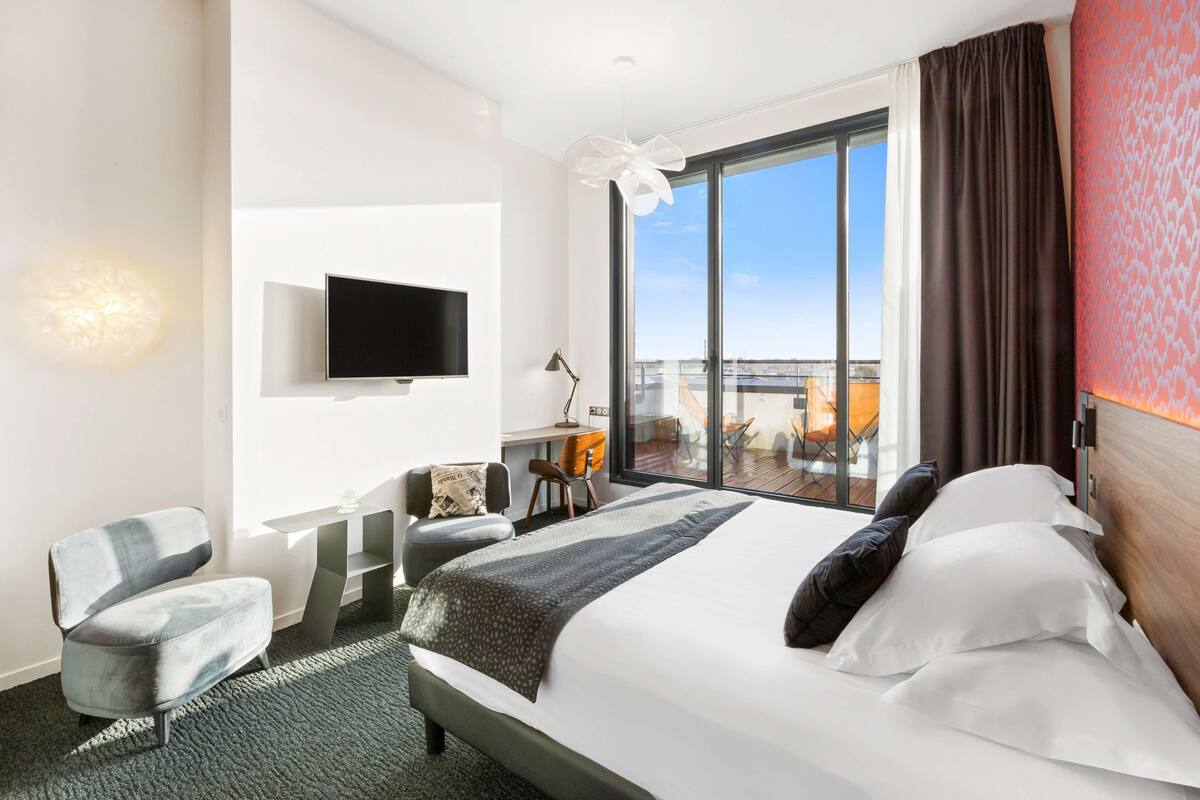 Quarto de hotel moderno com uma cama grande, televisão de tela plana na parede, escrivaninha com cadeira e varanda com vista para a cidade.