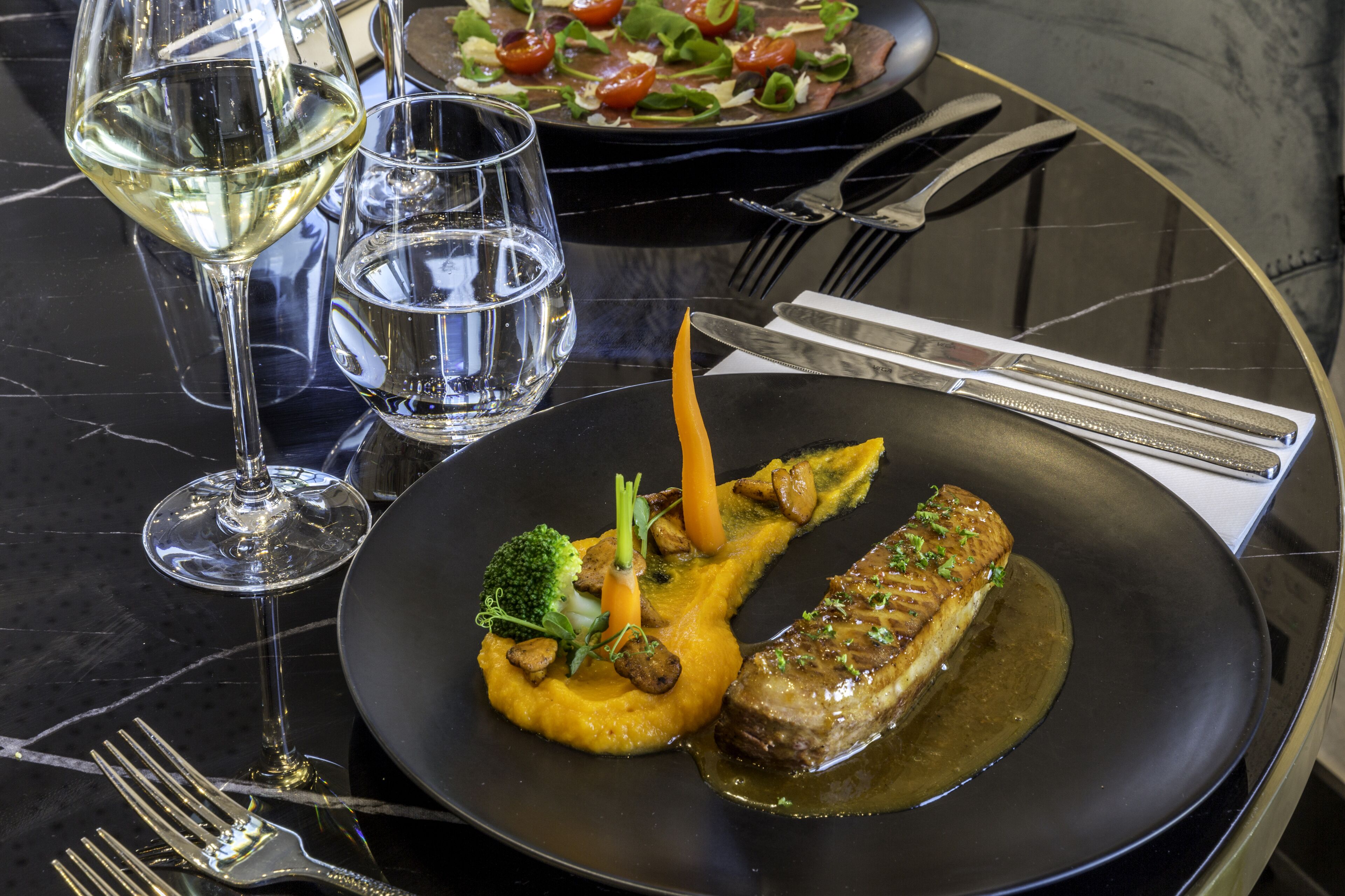A dining table with a black plate containing a fish fillet, vegetables, and a sauce. There are also two glasses of water and a fork on the table.