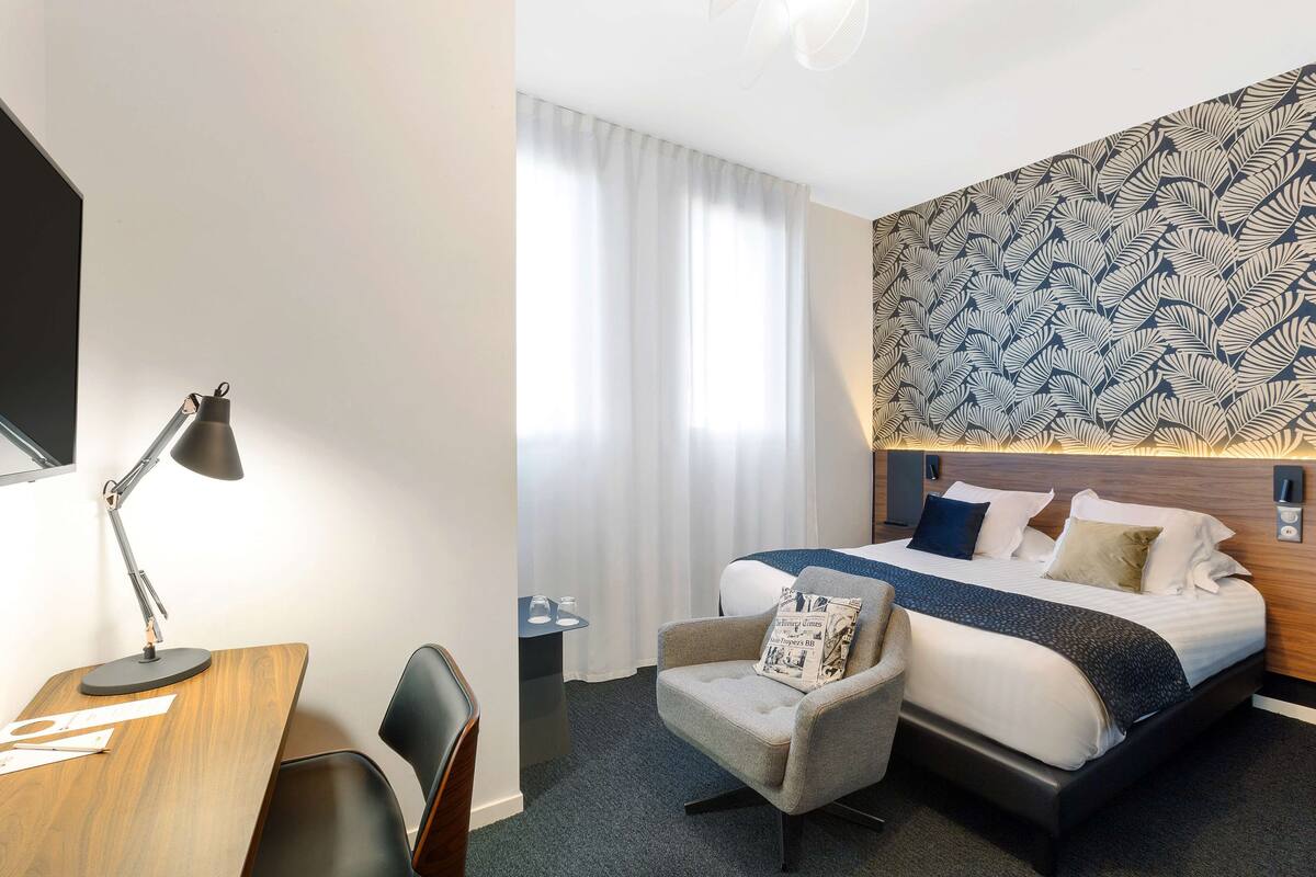 Quarto de hotel com escrivaninha de madeira, uma cadeira, cama com cabeceira estampada e uma janela com cortinas.