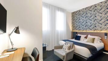 Una habitación de hotel con un escritorio de madera, una silla, una cama con cabecera estampada y una ventana con cortinas.