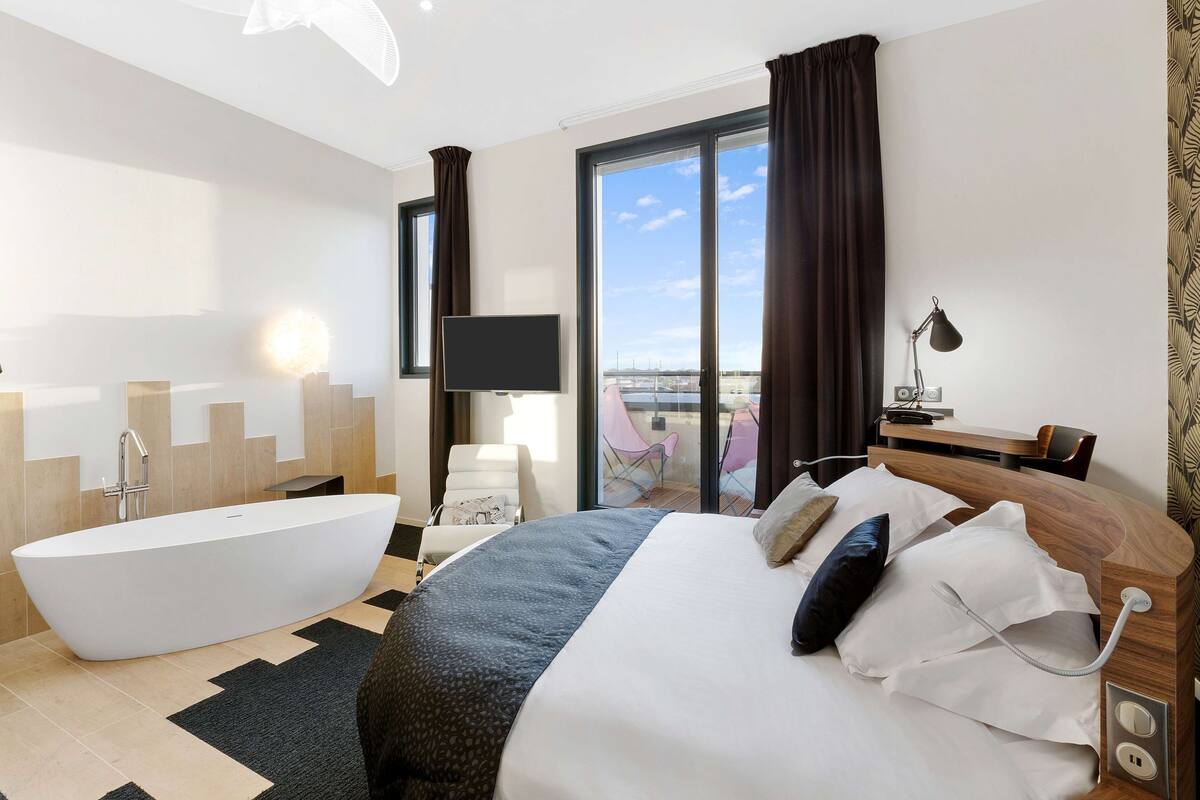 Quarto de hotel moderno com uma cama grande, banheira, televisão de tela plana e varanda com vista.