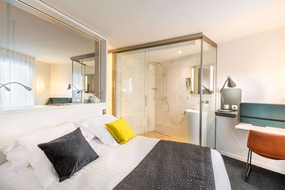 Quarto de hotel com cama, escrivaninha, cadeira, chuveiro e um espelho grande.