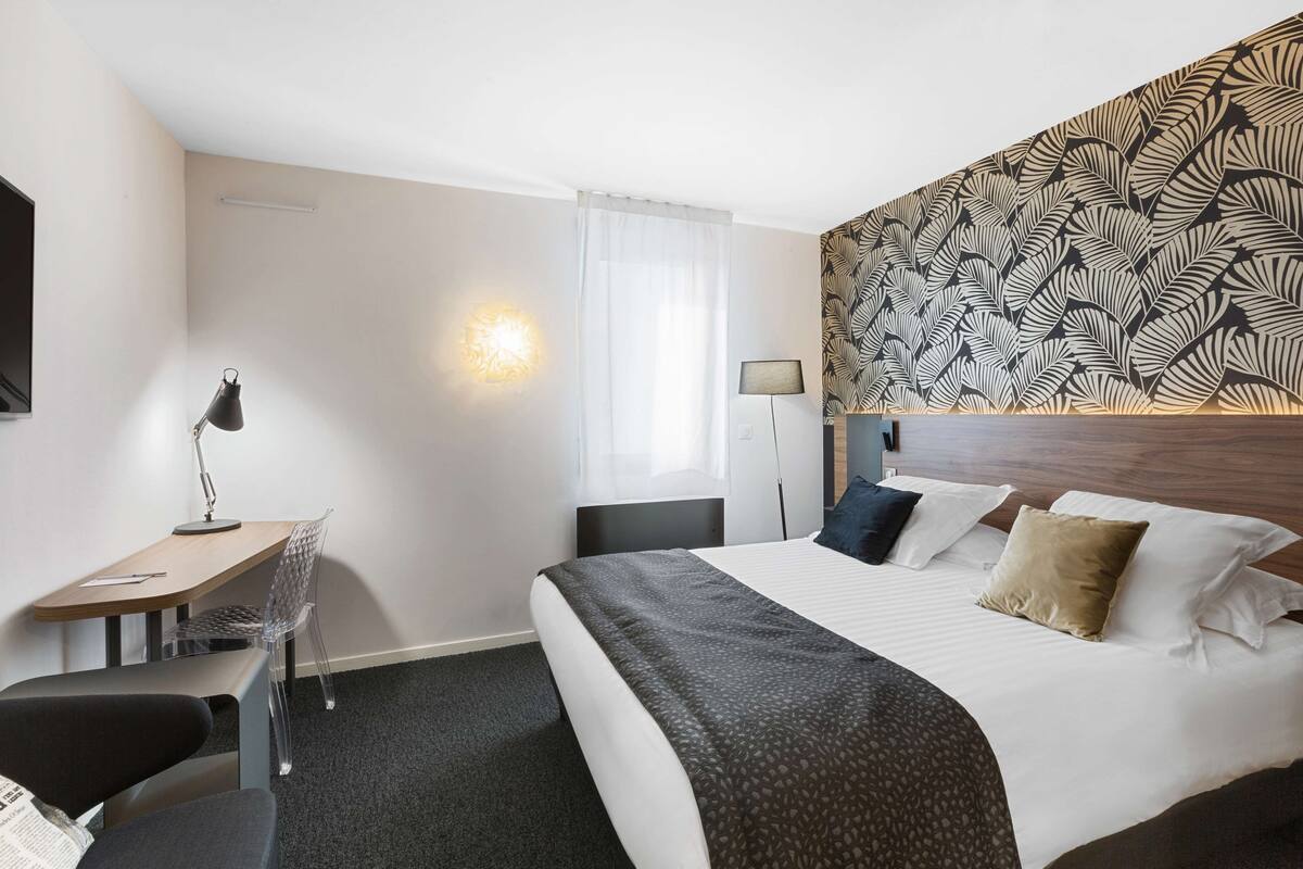 Quarto de hotel com uma cama, escrivaninha, cadeira, abajur e parede com padrão de folhagens.