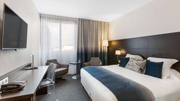 Habitación de hotel con una cama grande, un escritorio con silla, televisor, una mesita de noche con lámpara y ventana con cortinas.
