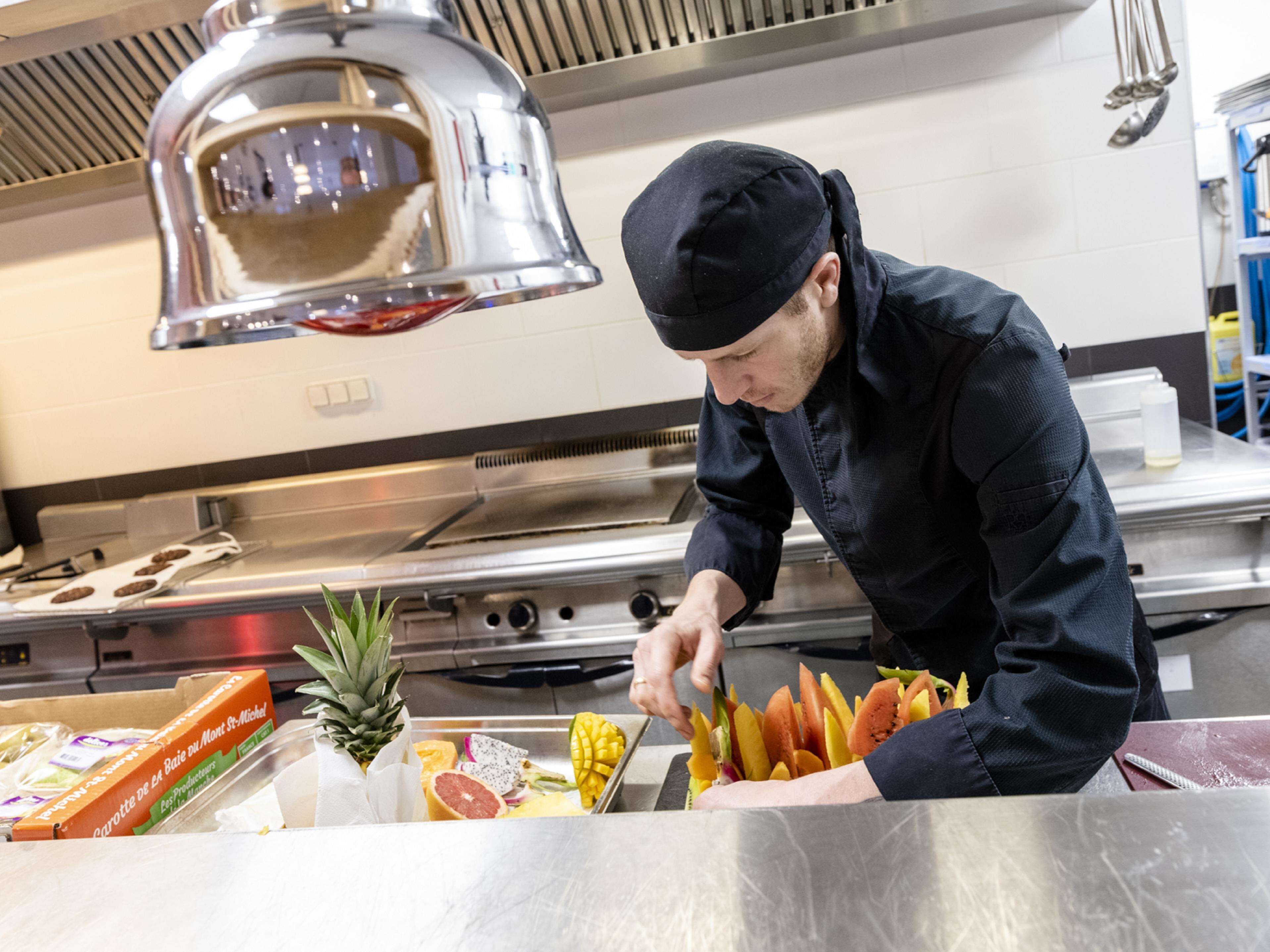 A chef preparing food in a professional kitchen with stainless steel appliances and a variety of fresh fruits and vegetables on the counter.