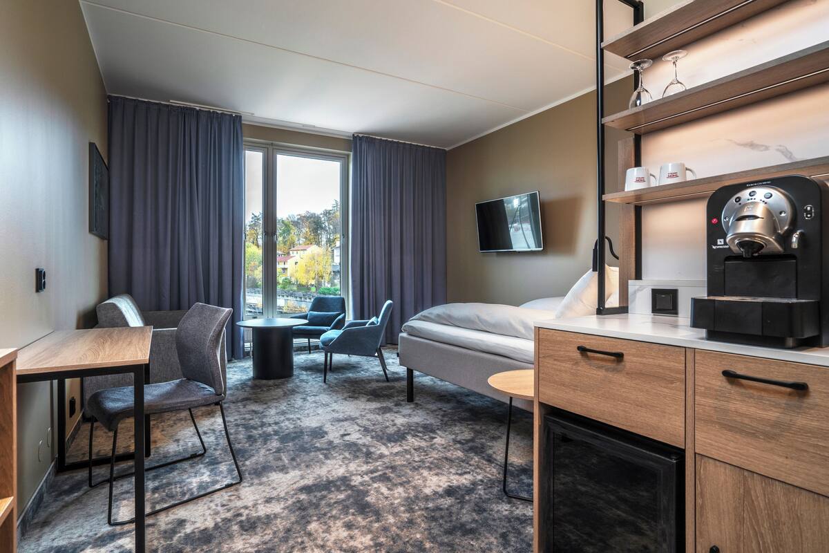A hotel room with a bed, a desk, two chairs, a coffee machine, a TV, and a window with curtains.