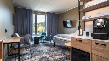 Habitación de hotel con cama, escritorio, dos sillas, cafetera, televisor y ventana con cortinas.