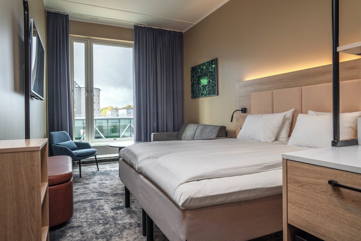 A hotel room with a large bed, a desk, a chair, a TV, and a view of the city through the window.