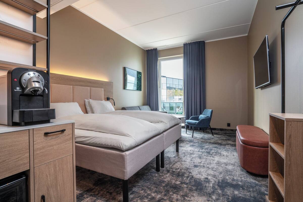 A hotel room with a bed, a coffee machine, a TV, a chair, and a window with curtains.