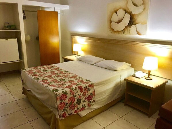 A hotel room with a bed, two bedside lamps, a nightstand, a wooden wardrobe, and a wall-mounted artwork.