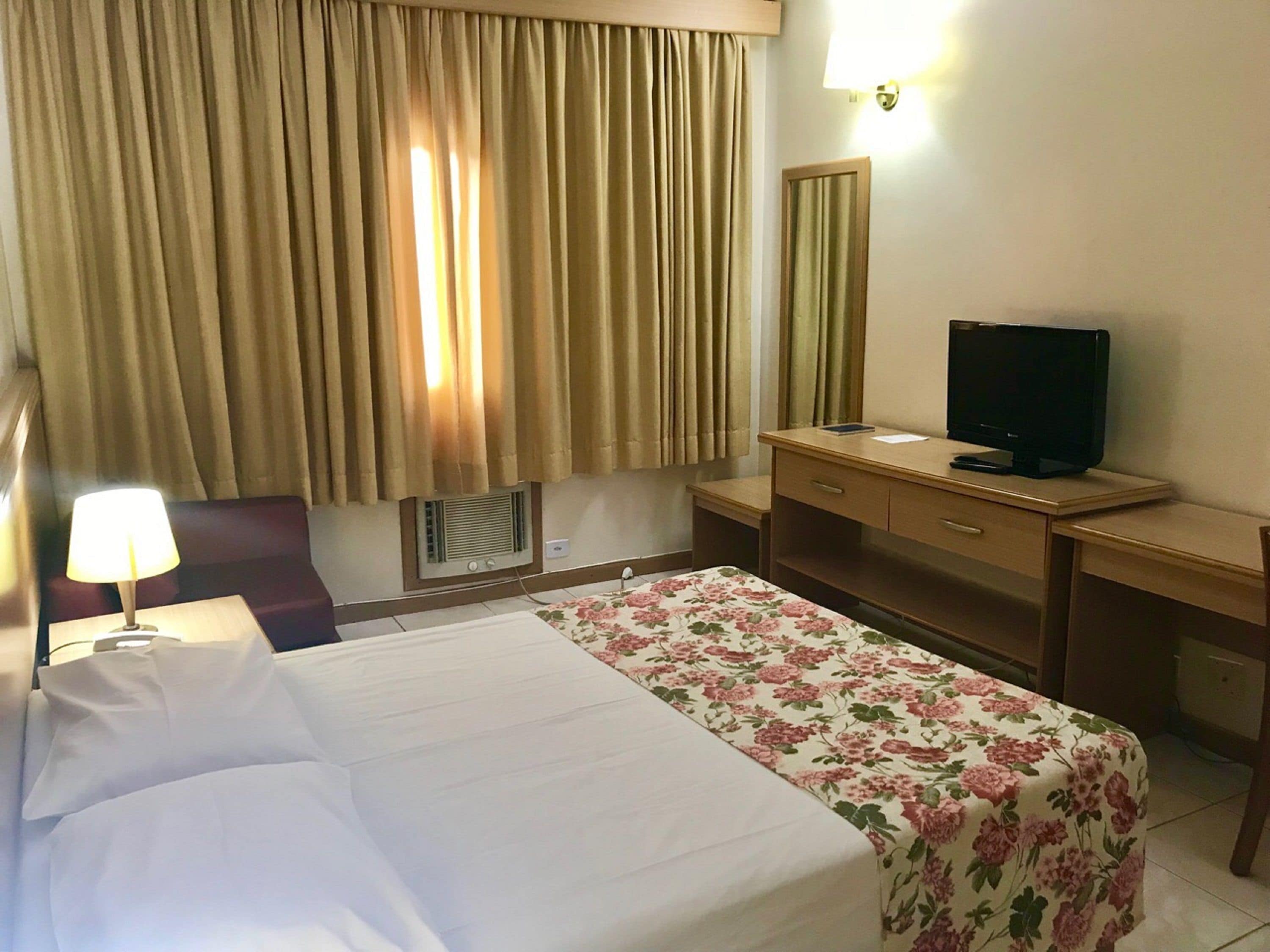Ein Hotelzimmer mit Bett, Schreibtisch, Fernseher und einem fensterbepflanzten Fenster.