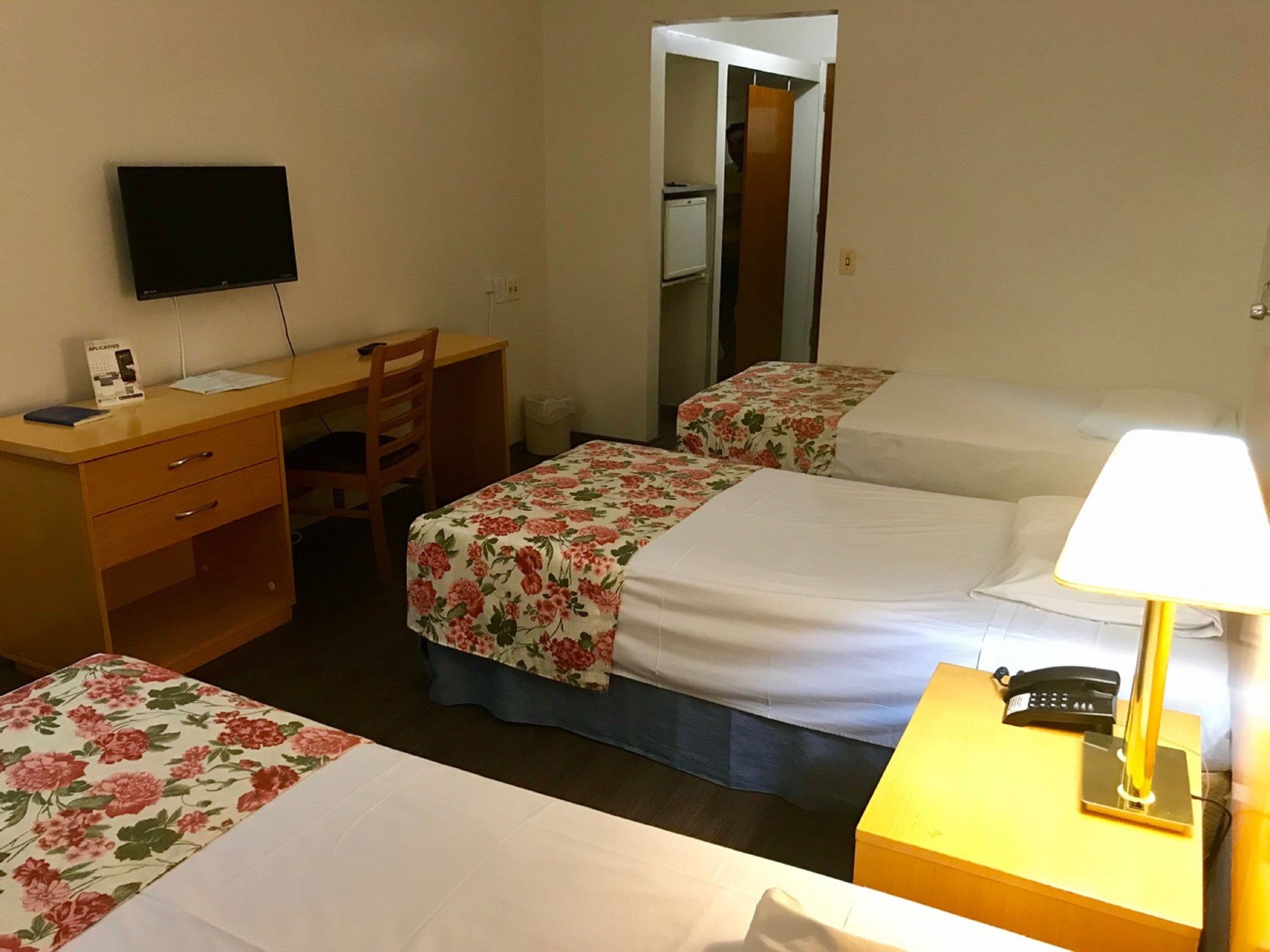 A hotel room with a desk, a chair, a bed with a floral bedspread, a bedside table with a lamp, and a mirror.