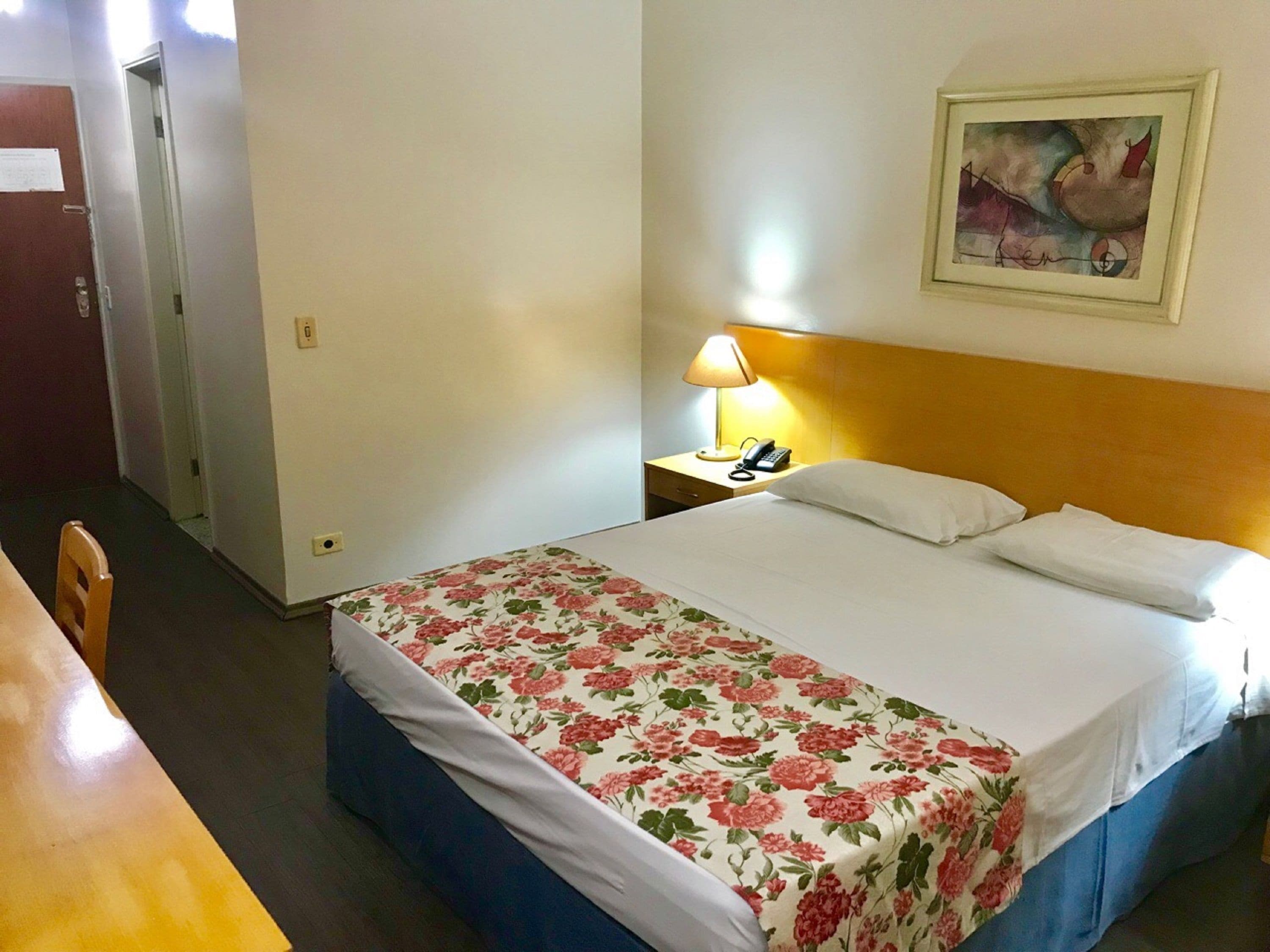Una habitación de hotel con cama, mesita de noche, lámpara, silla y un cuadro en la pared.
