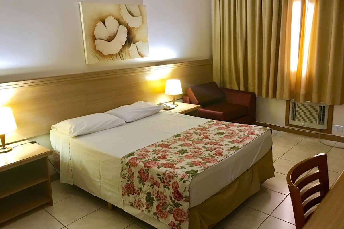 Een hotelkamer met een bed, een bureau, een stoel, een lamp, een schilderij aan de muur en een raam met gordijnen.