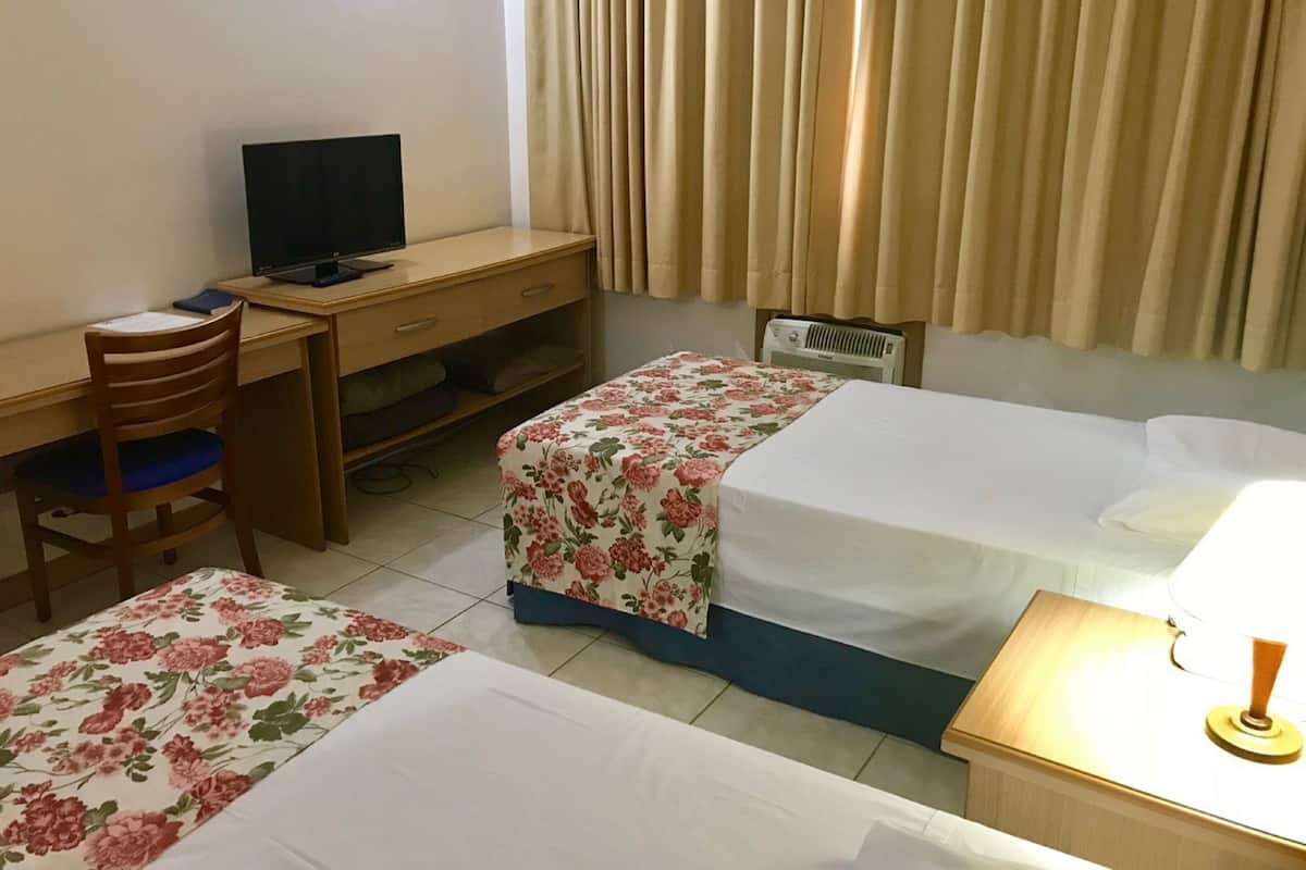 Een hotelkamer met een bureau, twee bedden, een stoel, een lamp en een raam met gordijnen.