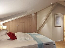 Un dormitorio con techo inclinado, cama con ropa de cama blanca y almohadas rojas, manta azul claro, armarios de madera y una obra de arte enmarcada en la pared.