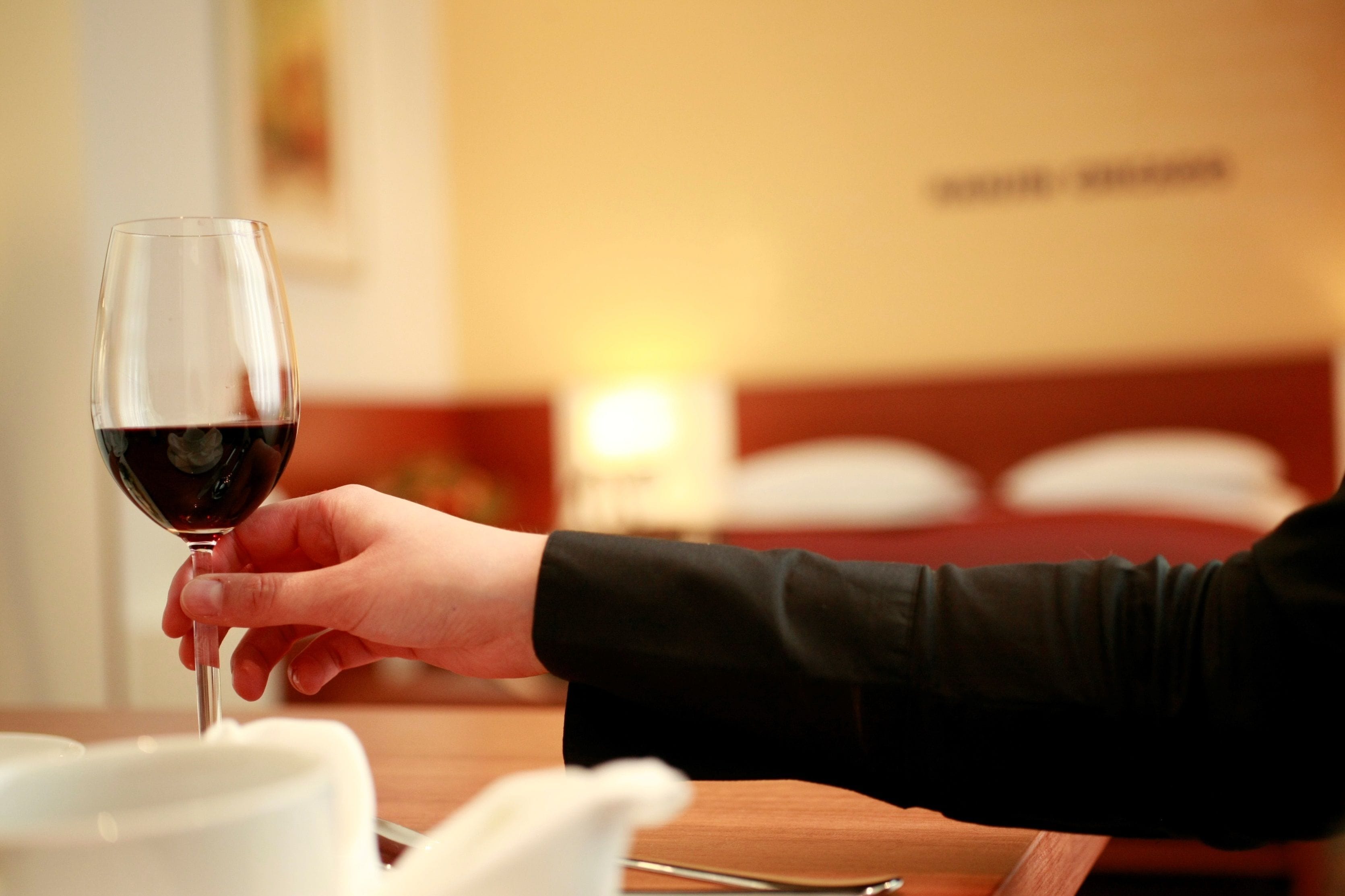 A hand holding a wine glass with a red liquid, a wooden table, and a bed with pillows in the background.