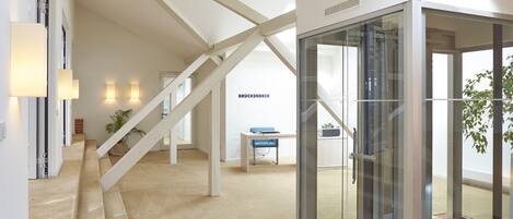 Un intérieur moderne avec un ascenseur en verre, un escalier et un comptoir d’accueil.