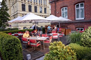 Un café al aire libre con mesas y sillas, rodeado de follaje y un edificio al fondo.