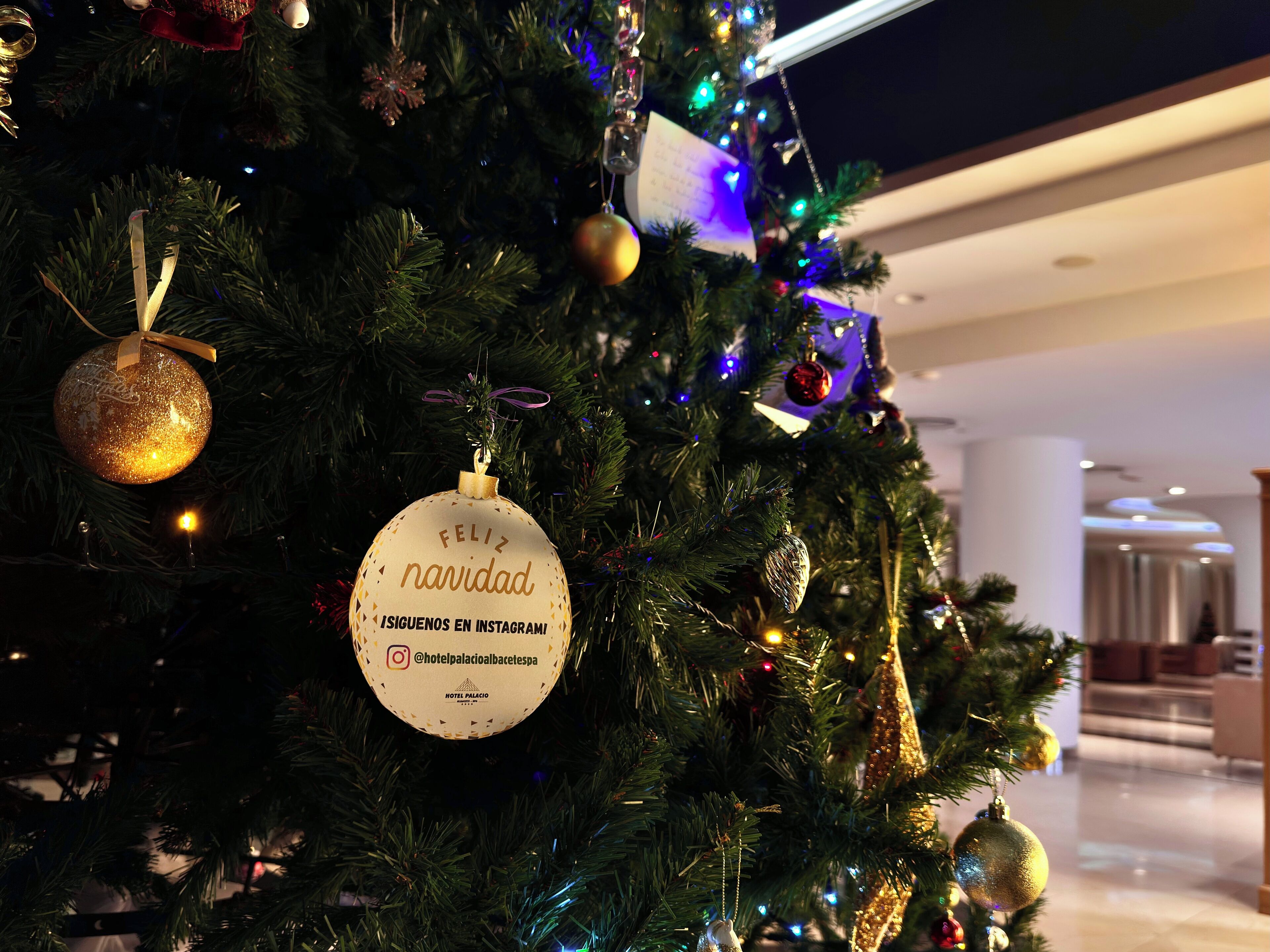 Un árbol de Navidad adornado con adornos y una tarjeta que dice "Feliz Navidad" y "Hotel Palacio Albacete SPA".