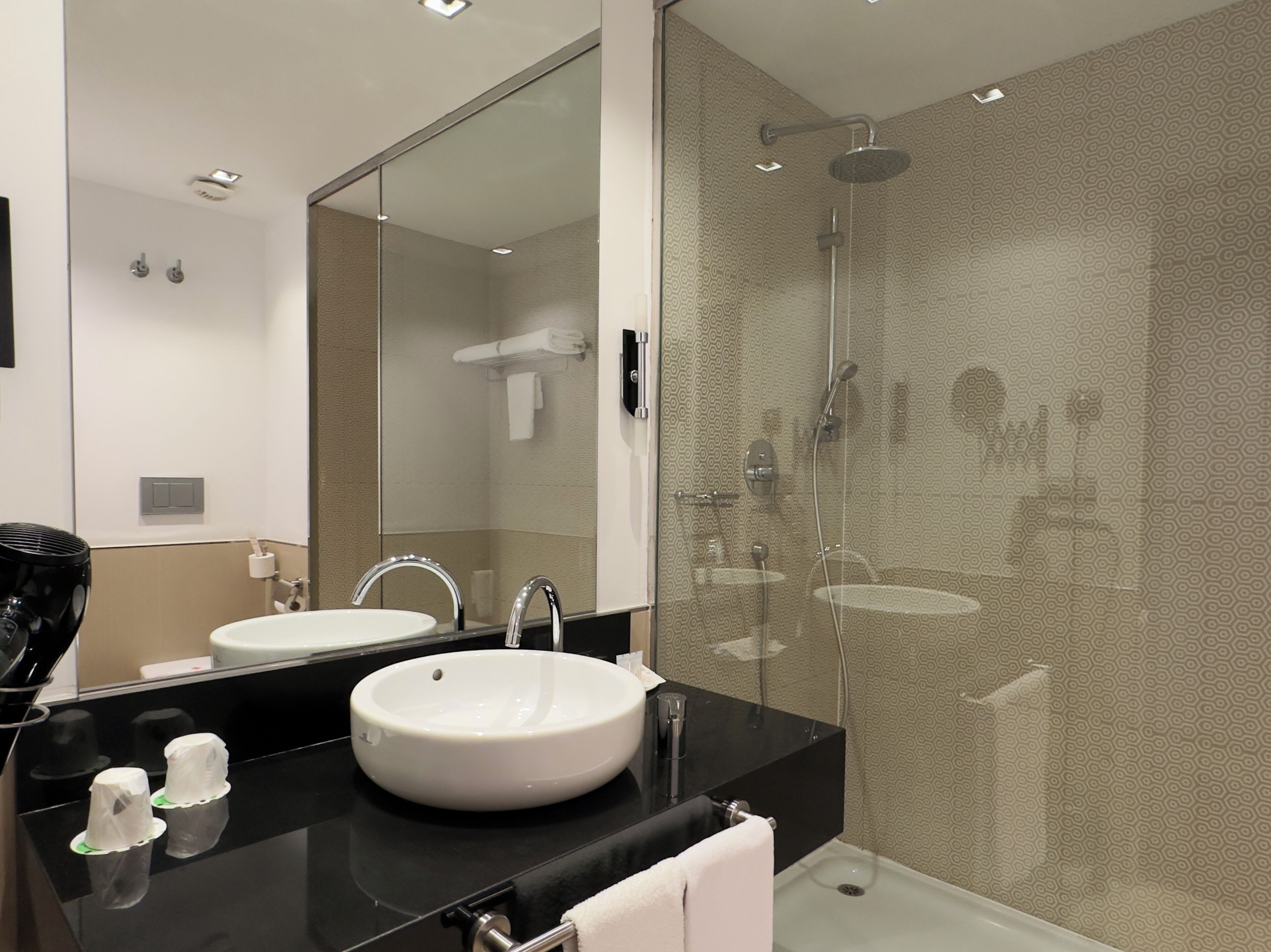 A modern bathroom with a large mirror, a white sink, a black countertop, and a glass-enclosed shower area.