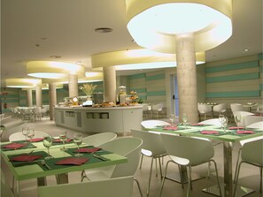 A modern dining area with multiple tables and chairs, a central counter with food display, and a ceiling with circular light fixtures. - Hotel Palacio Albacete & SPA (Albacete)