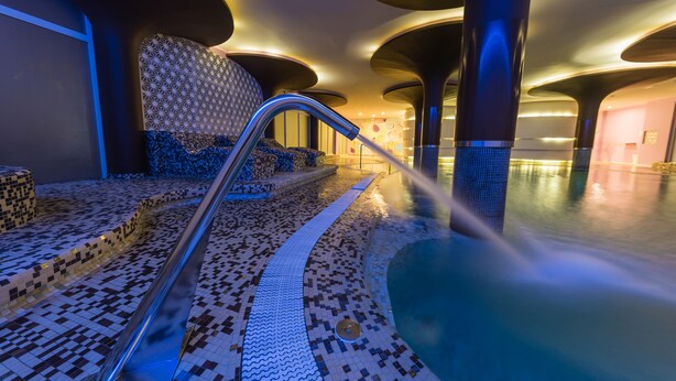 A modern indoor pool area with a mosaic tile floor, a water feature, and unique ceiling architecture. - Hotel Palacio Albacete & SPA (Albacete)