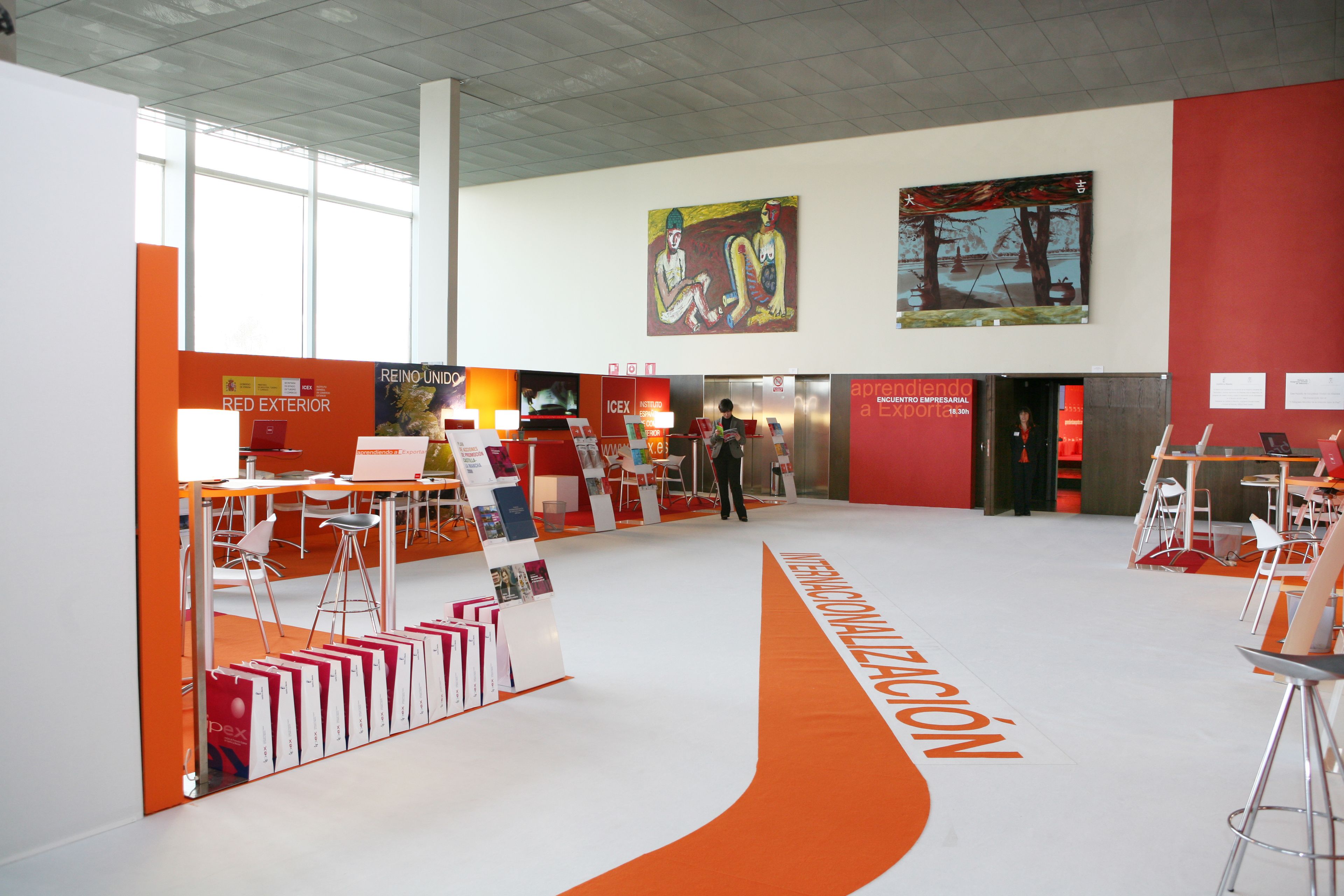 Un amplio salón de exposiciones con paredes blancas y anaranjadas, que cuenta con un pasillo central y varias mesas y sillas.