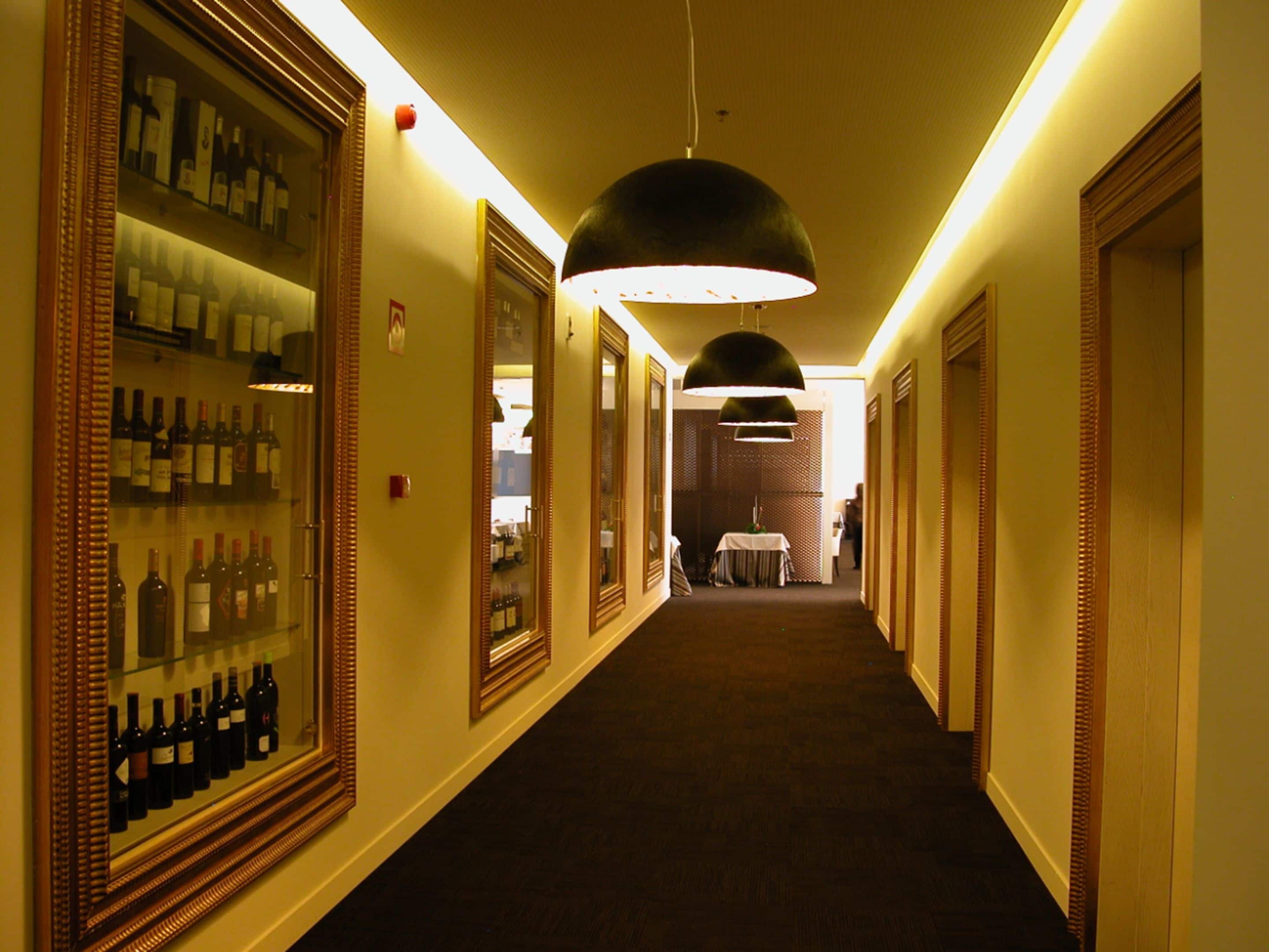 A hotel corridor with framed wine bottles on the left, a table with a chair in the center, and a hanging light fixture.