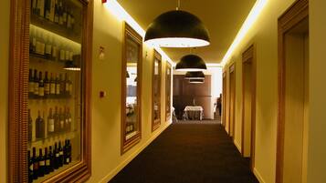 A hotel corridor with framed wine bottles on the left, a table with a chair in the center, and a hanging light fixture.