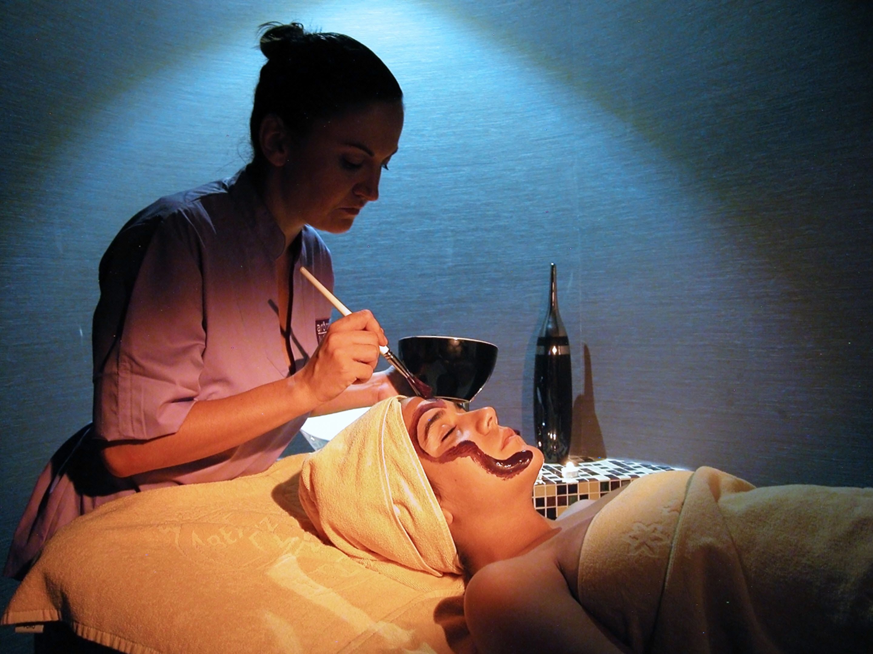 A person receiving a facial treatment in a dimly lit room with a tiled wall and a bottle on a small table.