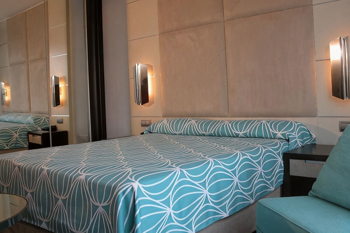 A hotel room with a large bed, a sitting area, a mirror, and wall-mounted lights.