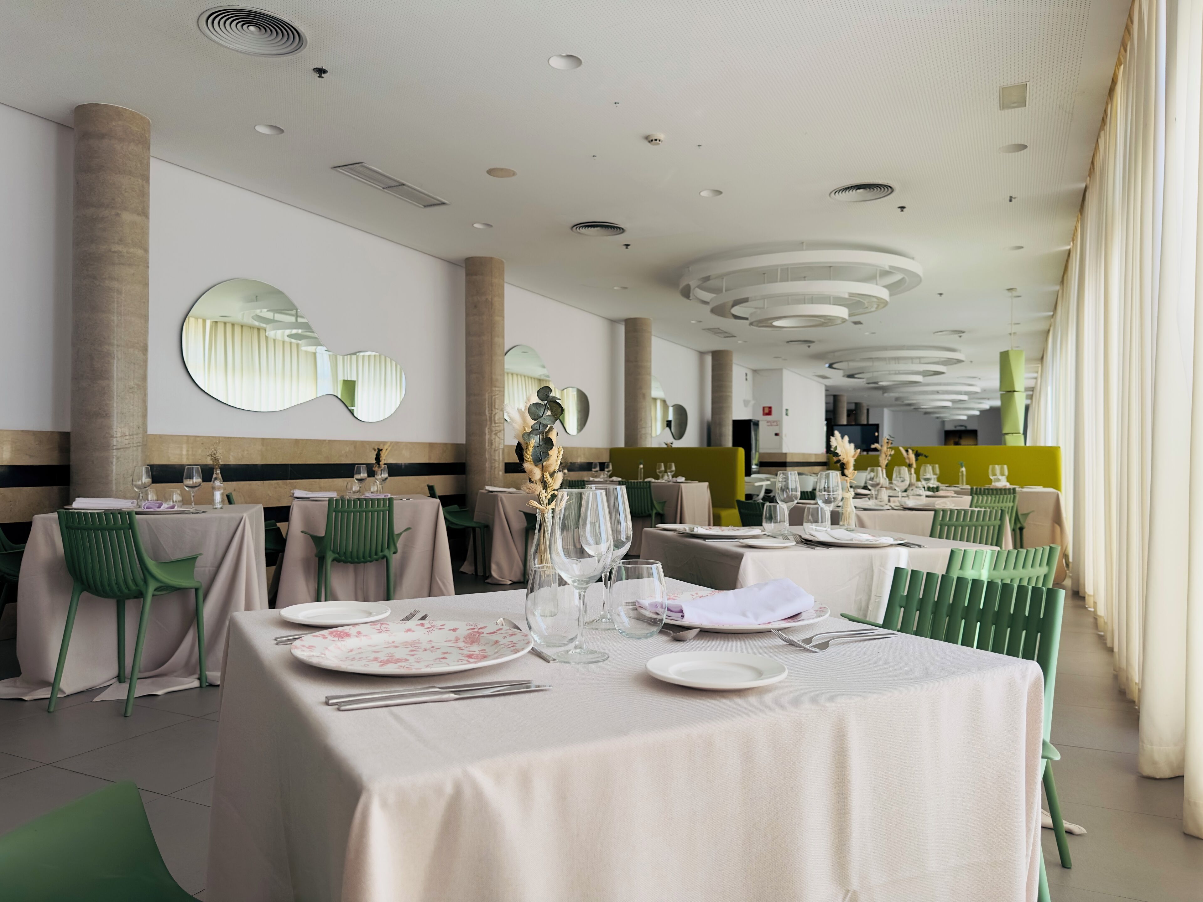 A restaurant with round tables, green chairs, and a modern ceiling design.