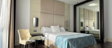 A hotel room with a bed, a chair, a small table, a mirror, and a wall-mounted TV.